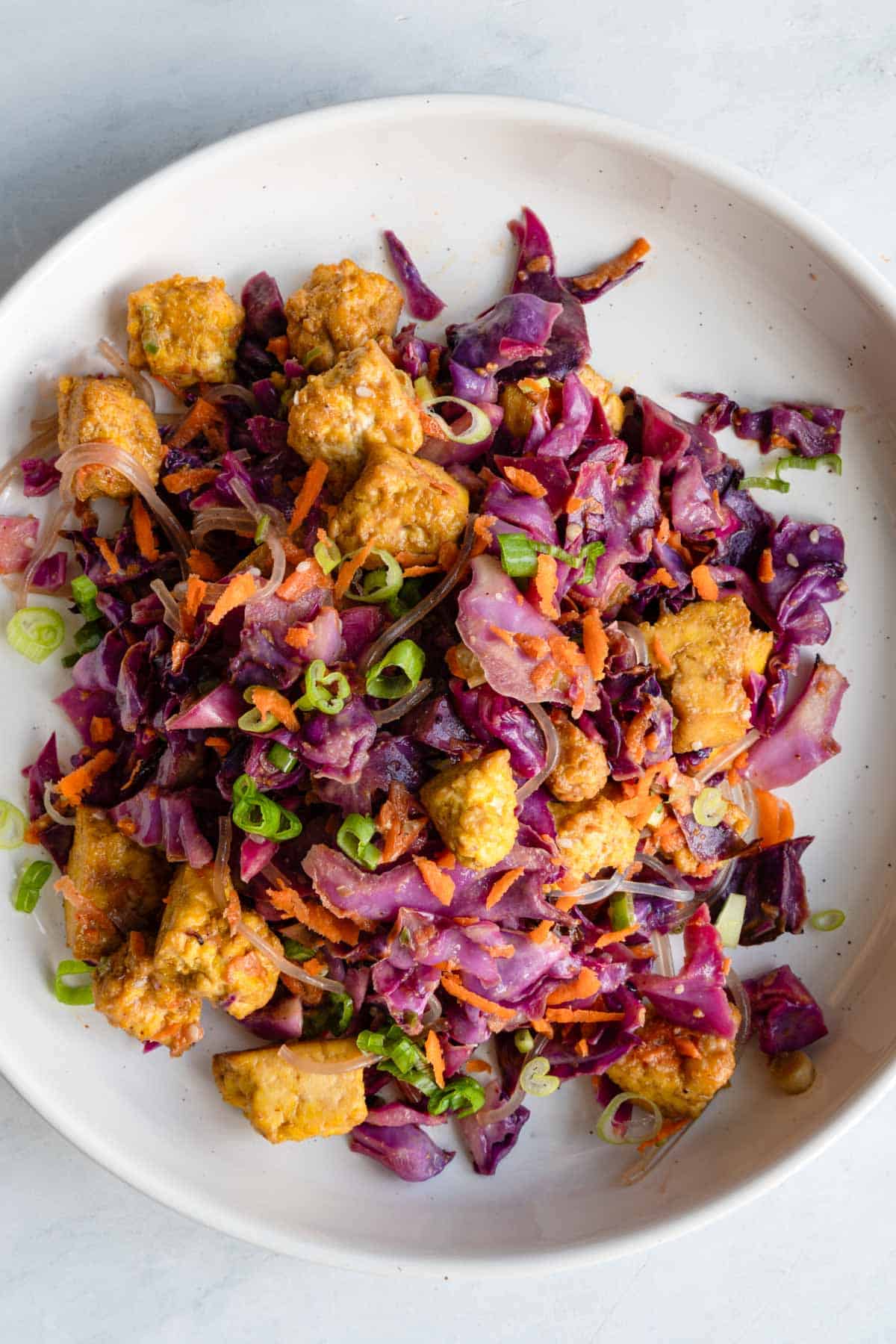 Tofu Salad with roasted tofu and red cabbage, grated carrots, chopped scallions, sesame seeds, glass noodles, and Asian dressing in a white bowl.