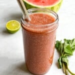 Watermelon juice with chia seeds in a glass with a metal straw. Plus, one-half watermelon behind, one-half lime, and fresh mint.