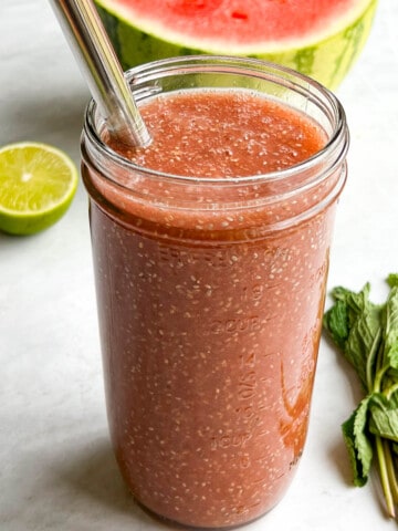 Watermelon juice with chia seeds in a glass with a metal straw. Plus, one-half watermelon behind, one-half lime, and fresh mint.