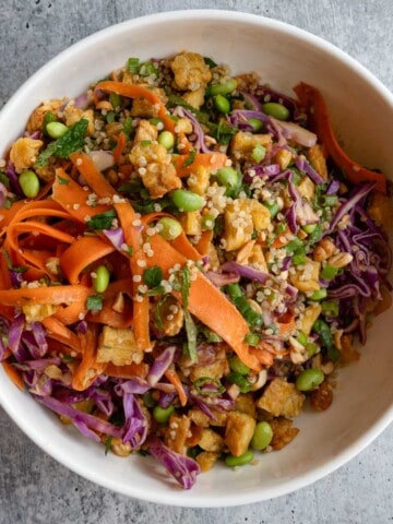 Baked tempeh cubes, shredded red cabbage, ribbon carrots, edamame, chopped green onion, quinoa, chopped mint, and peanut dressing in a white bowl.