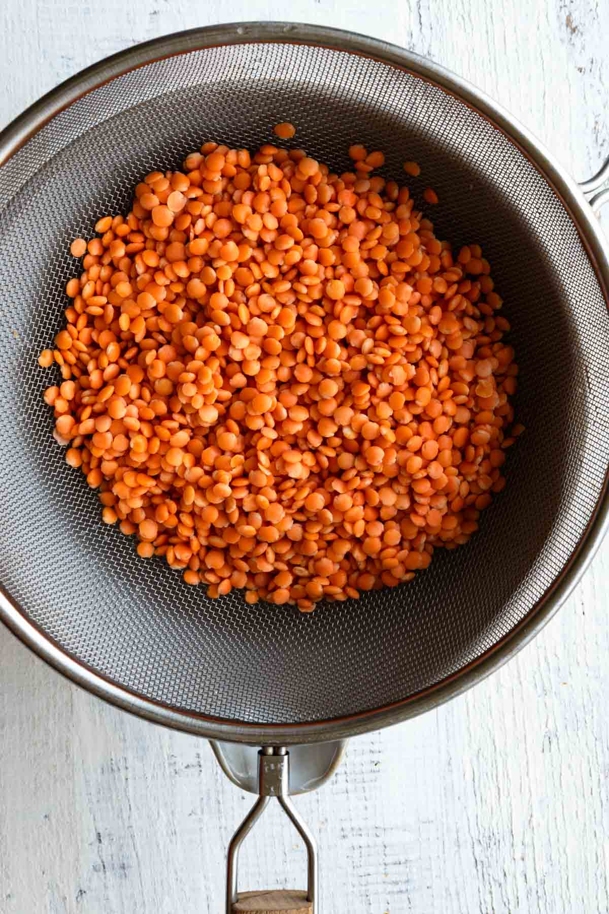 Washed red lentils draining in a silver sieve over a white bowl.