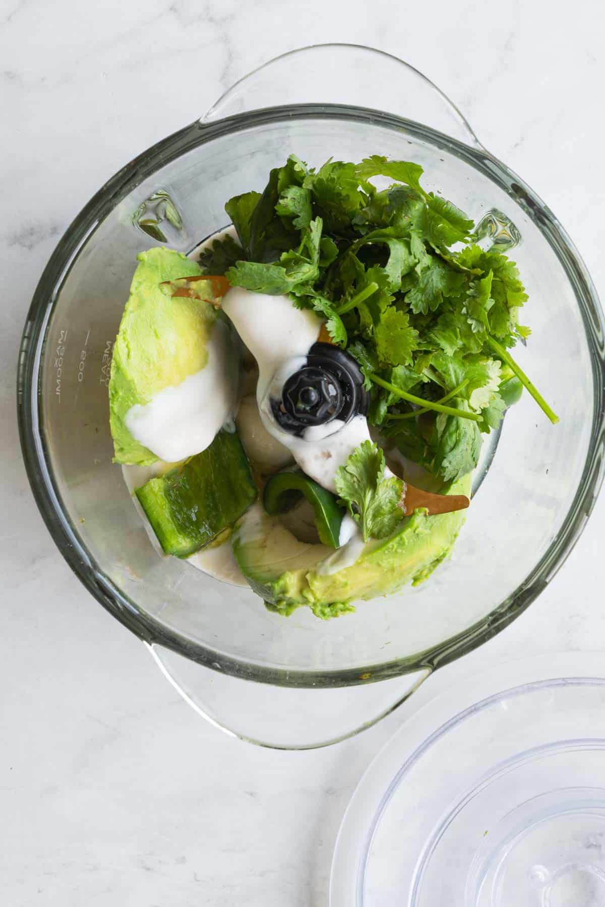 Avocado, cilantro, chopped jalapeño, garlic and vegan yogurt in a glass canister of a food processor.