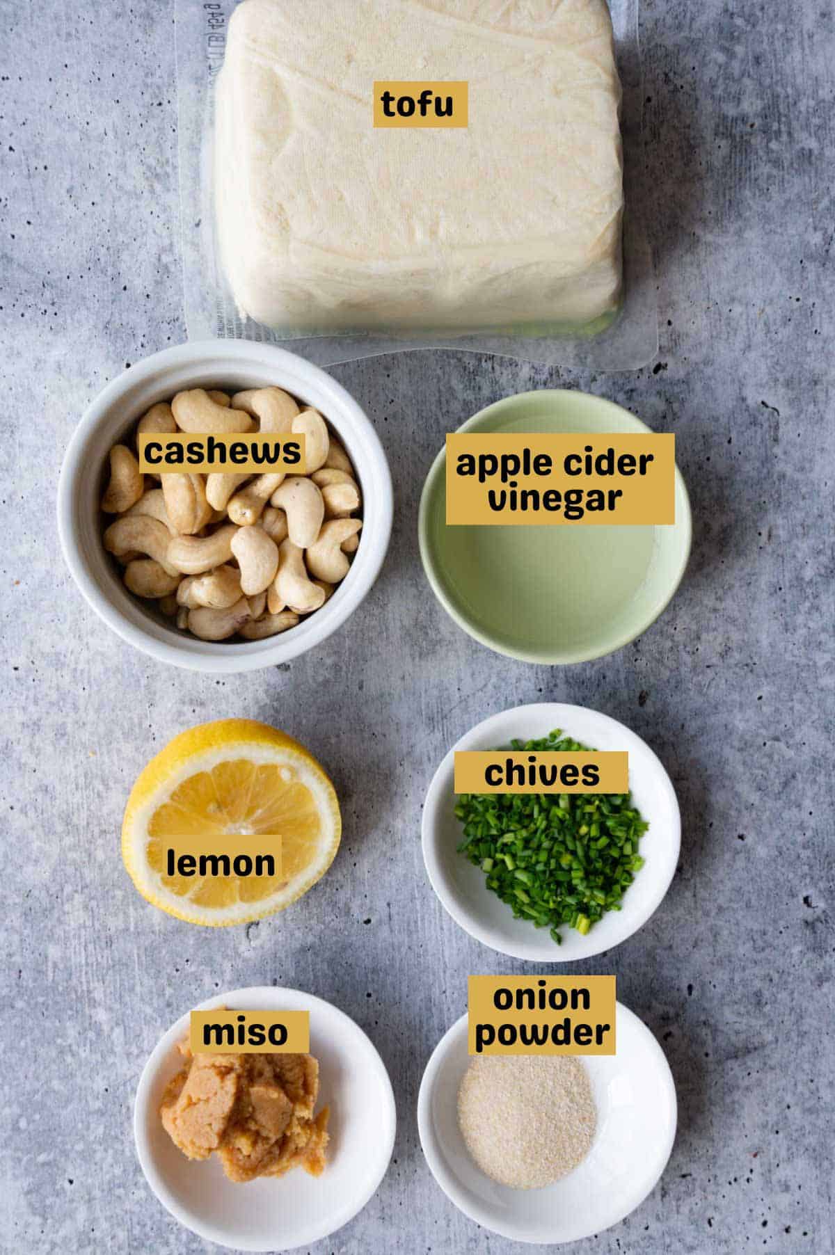 Tofu, cashews, apple cider vinegar, lemon juice, miso, onion powder, lemon, and miso on a gray backdrop.