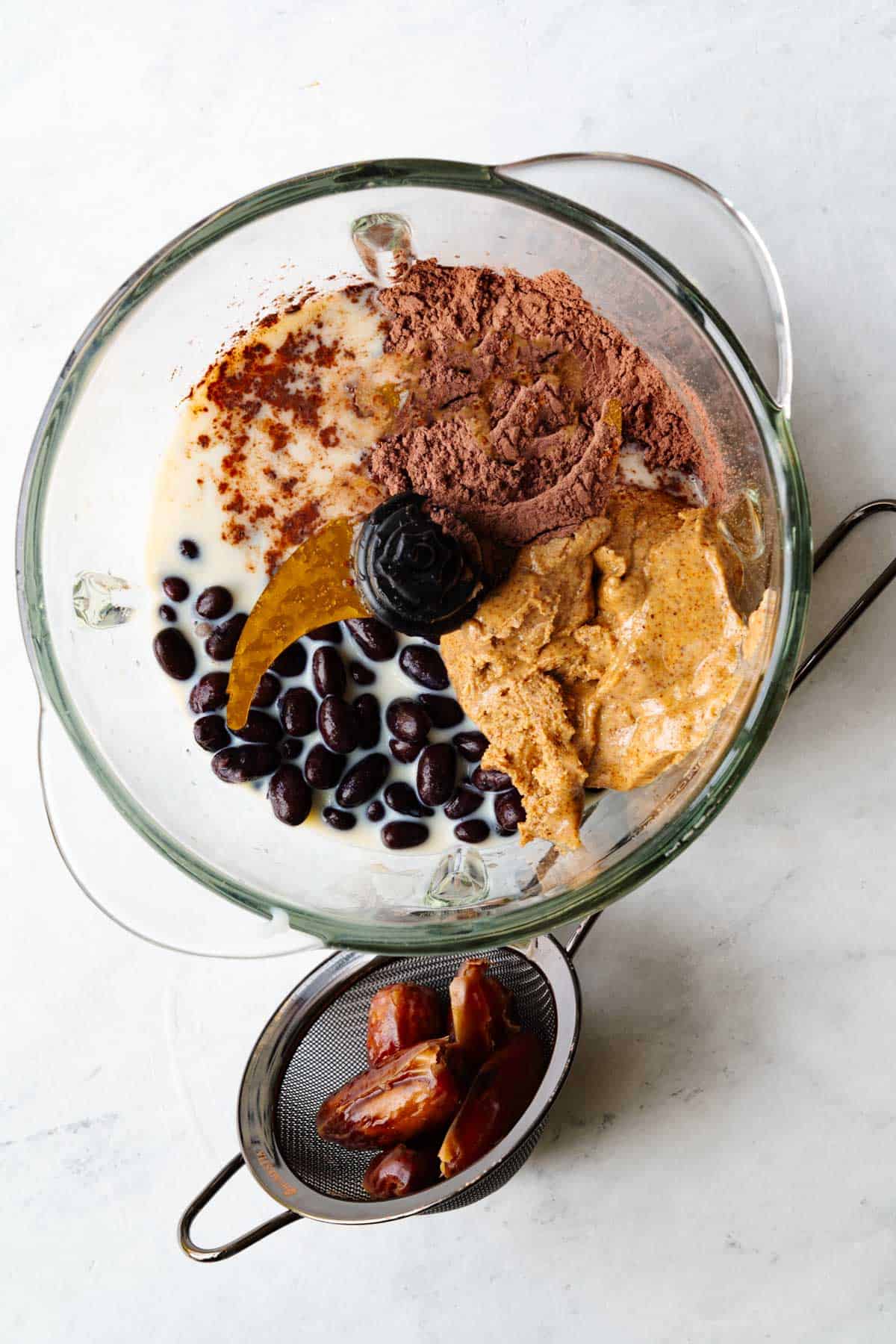 Almond butter, nondairy milk, black beans, cocoa powder, and vanilla extract in the glass canister of a food processor next to a mesh sieve with pitted dates.