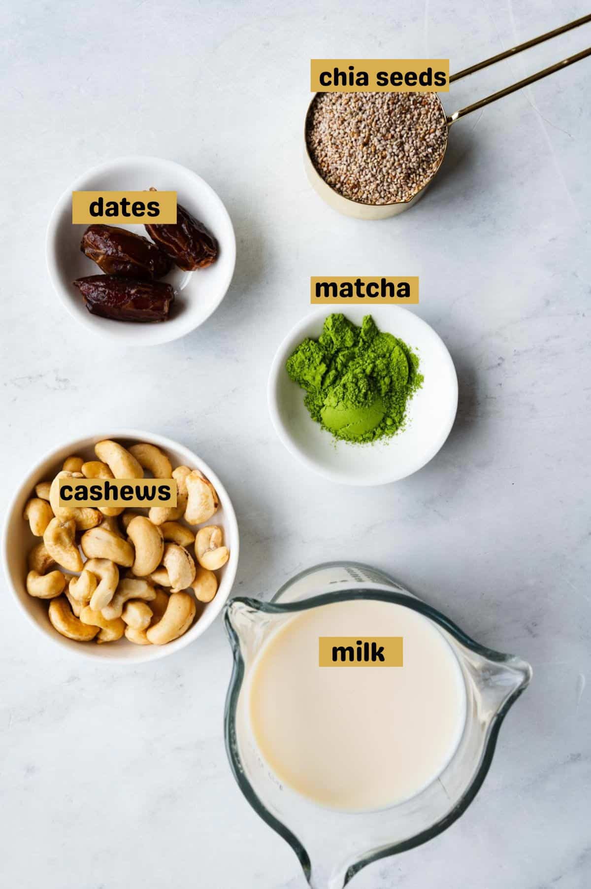 Chia seeds in a gold measuring cup, dates in a small white bowl, matcha powder and cashews in small white bowls, and milk in a glass beaker.