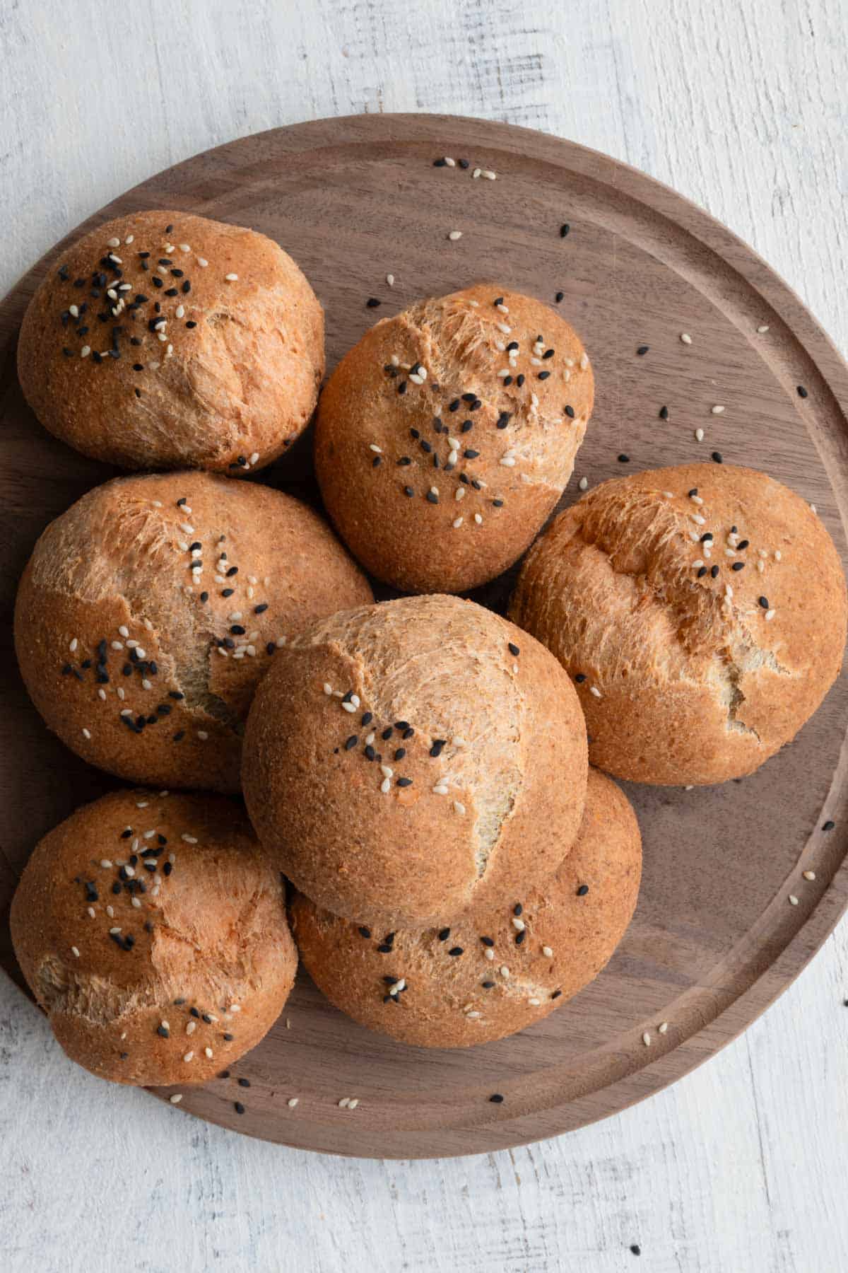 Seven red lentil gluten-free rolls with black and white sesame seeds on top on a round wooden cutting board.