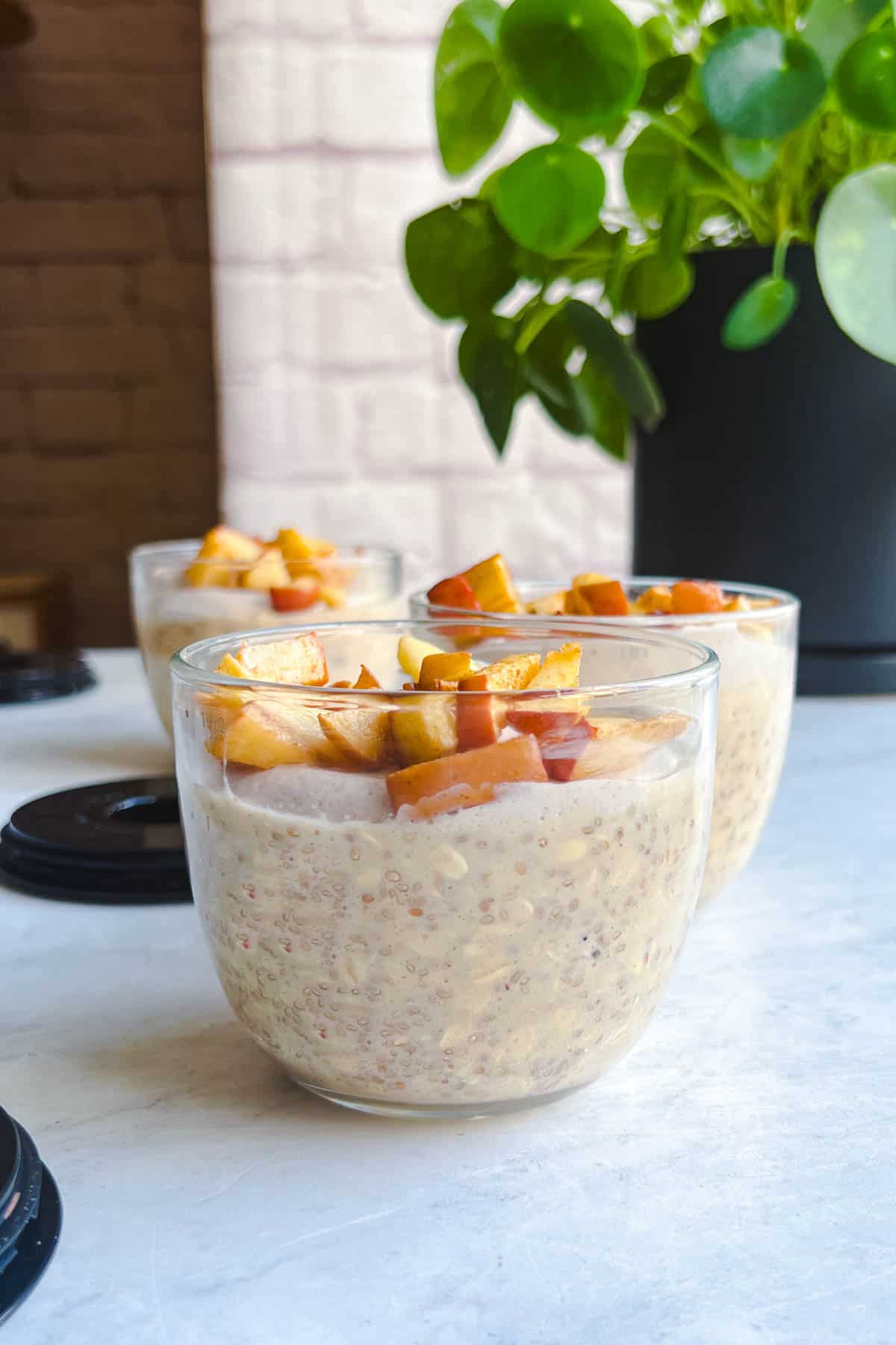 Apple cinnamon overnight oats with chia seeds in three glass jars topped with crispy baked apple in front of a green plant.