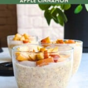 Apple cinnamon overnight oats with chia seeds in a three glass jars and topped with crispy baked apple sitting in front of a green plant.