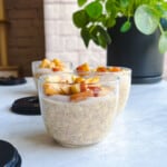 Apple cinnamon overnight oats with chia seeds in three glass jars topped with crispy baked apple in front of a green plant.
