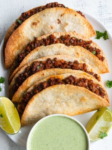 Walnut meat in tacos on a white plate with lime wedges and avocado sauce.