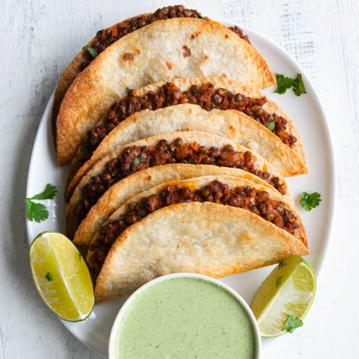 Walnut meat in tacos on a white plate with lime wedges and avocado sauce.