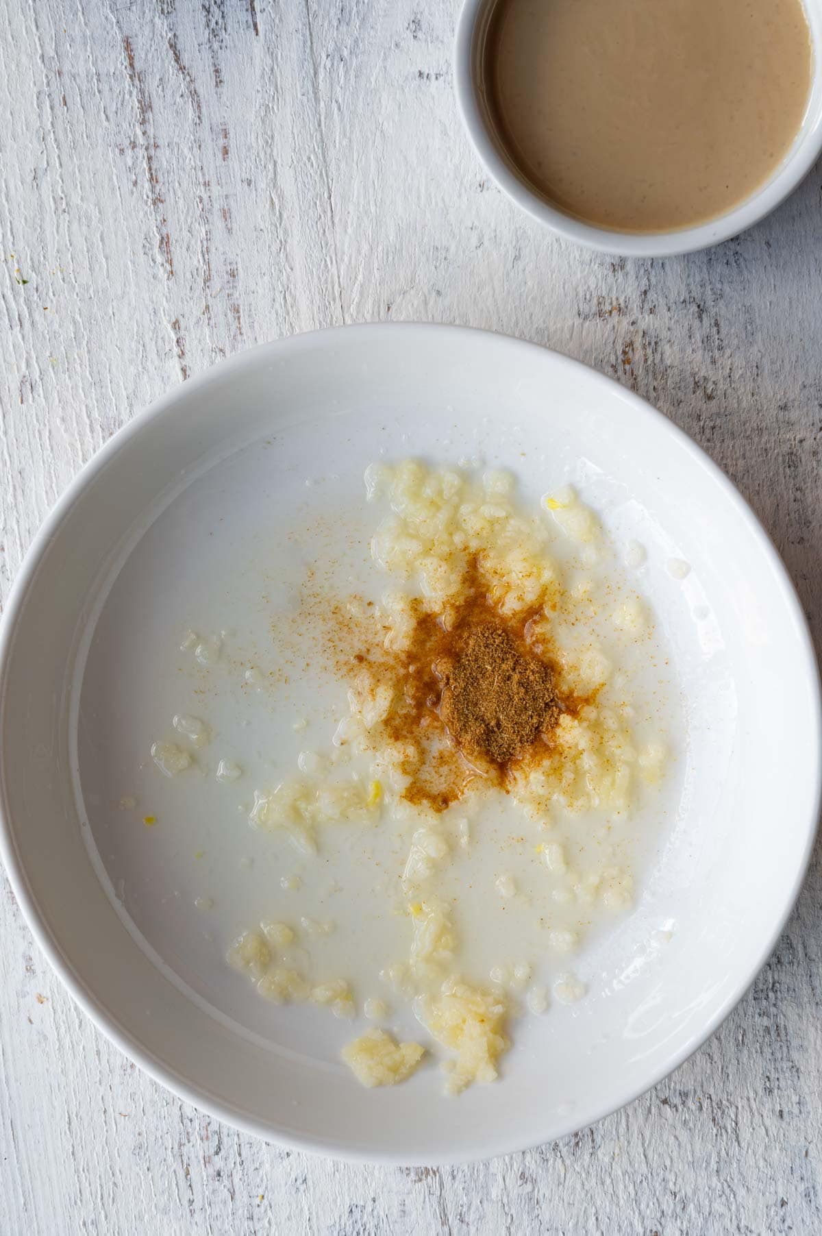 Minced garlic, lemon juice, and ground cumin in a white bowl next to a small white bowl with tahini.