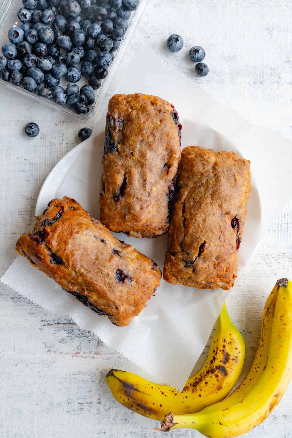 Three gluten-free vegan banana bread mini loaves on white parchment paper beside a box of blueberries and two bananas.