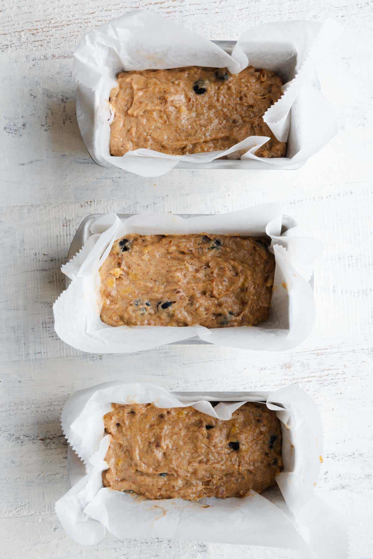 Yellow-brown batter with whole blueberries inside three white parchment-lined mini loaf pans.