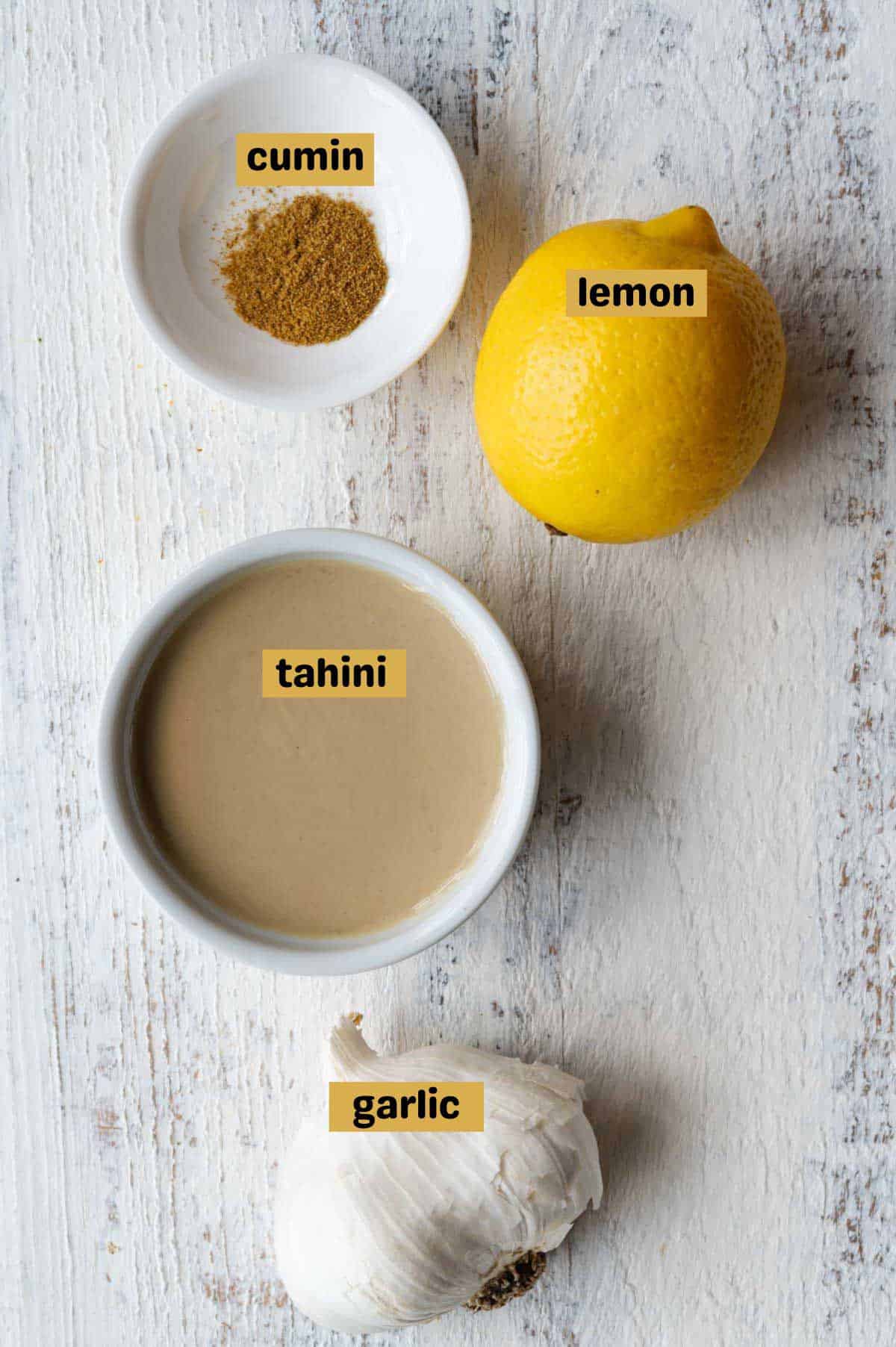 Cumin, lemon, tahini, and garlic on a white wooden backdrop.