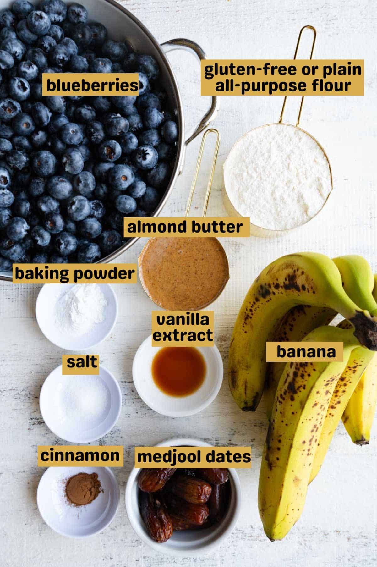 Blueberries, all-purpose flour, almond butter, maple syrup, salt, baking powder, bananas, and cinnamon in small bowls on a white backdrop.