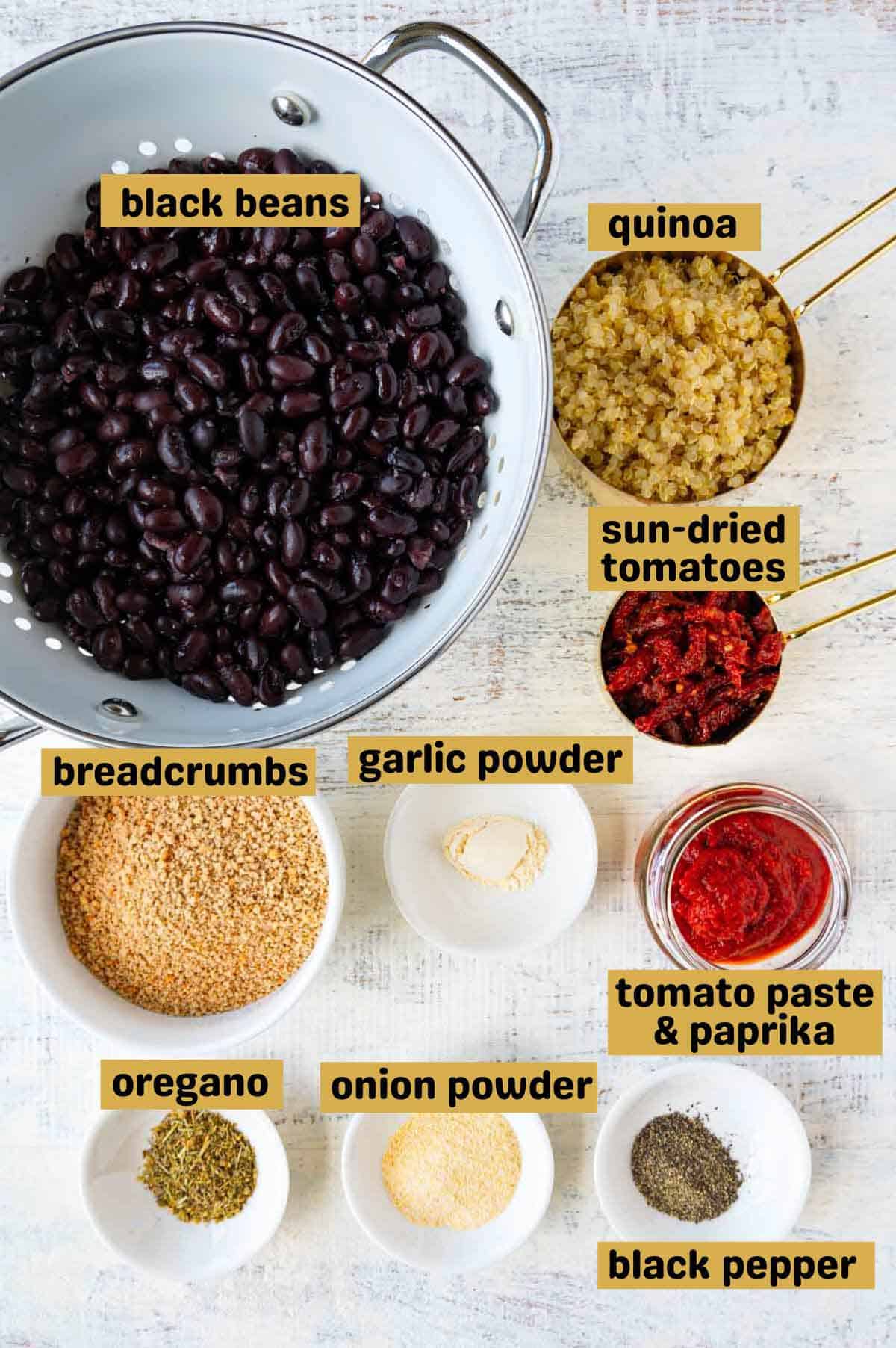 Black beans in a white colander, quinoa in a gold measuring cup, sun-dried tomatoes in a gold measuring cup, garlic powder, onion powder, oregano, breadcrumbs, and tomato paste in white bowls on a white backdrop.