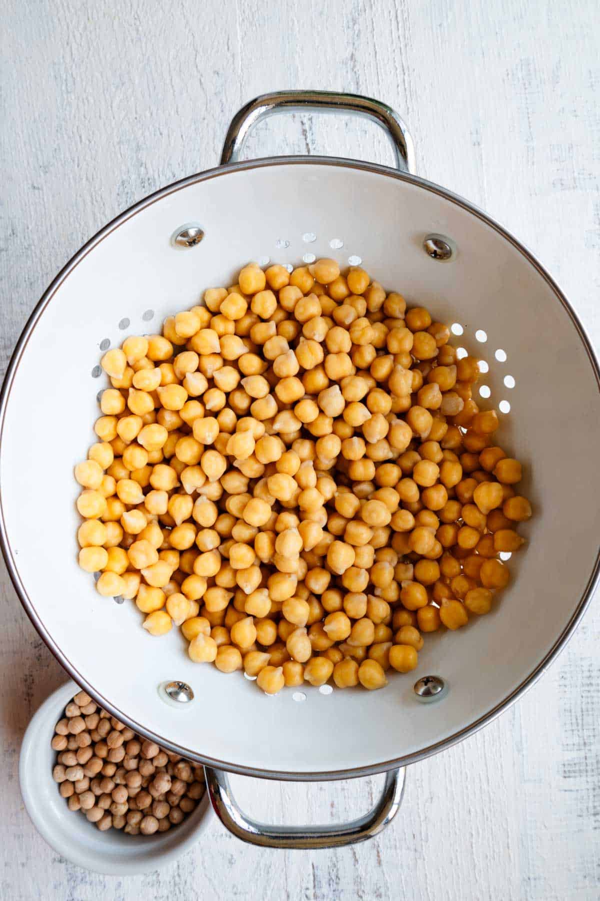 Soaked and drained chickpeas in a white colander beside a white bowl of dried chickpeas.