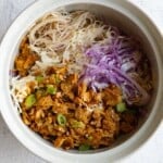 Cooked shredded tofu with green onion and sesame seeds, red cabbage, savoy cabbage, and brown rice vermicelli noodles in a white bowl.