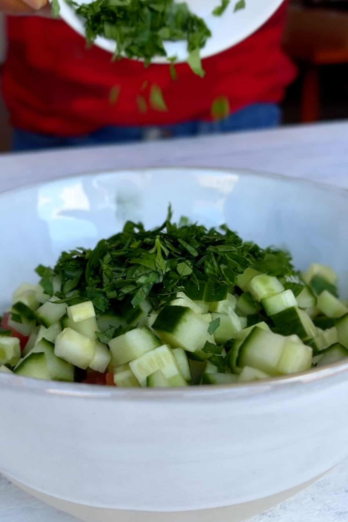 Pouring chopped fresh green herbs into a bowl of chopped cucumber and tomatoes.