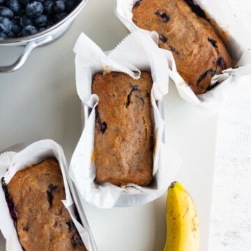 Three gluten-free vegan banana bread mini loaves in white parchment paper in ini loaf pans beside a colander of blueberries and one banana.