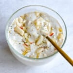 Homemade cashew yogurt in a glass bowl topped with chopped apples, drizzle agave, hemp hearts, and a gold spoon.