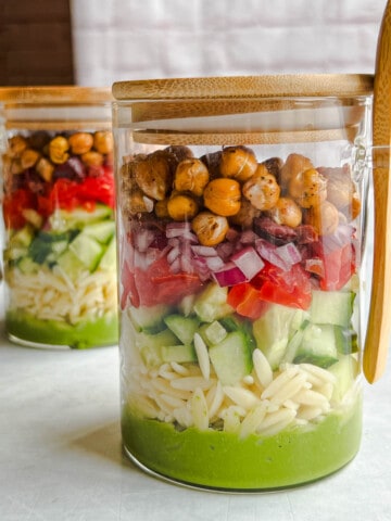 Two salad jars with green oil-free pesto on the bottom, orzo, chopped cucumber, chopped tomato, red onion, and oil-free roasted chickpeas, and a wooden spoon.
