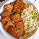 Golden brown crispy tempeh in marinade with noodles and sesame seeds and green onion in a white bowl.