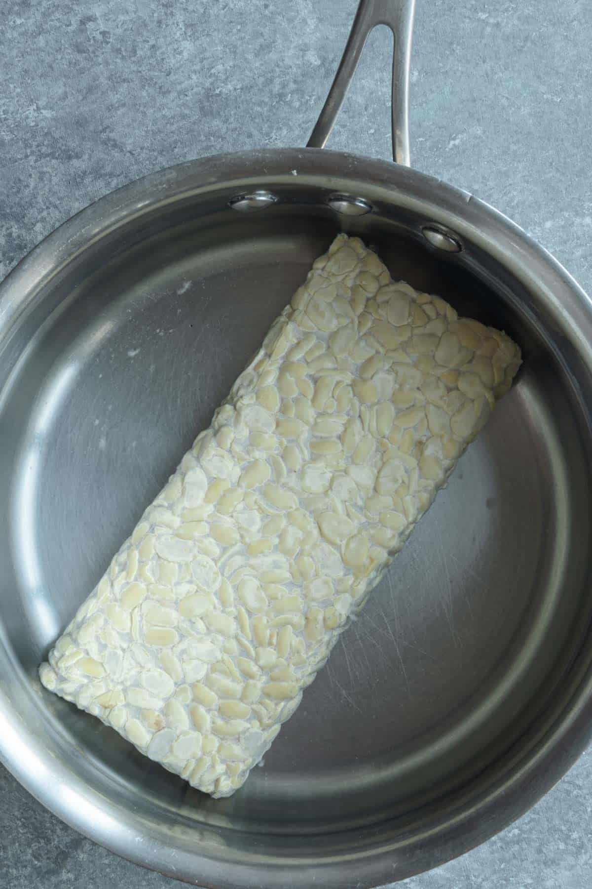 A block of tempeh simmering in a stainless steel saucepan.