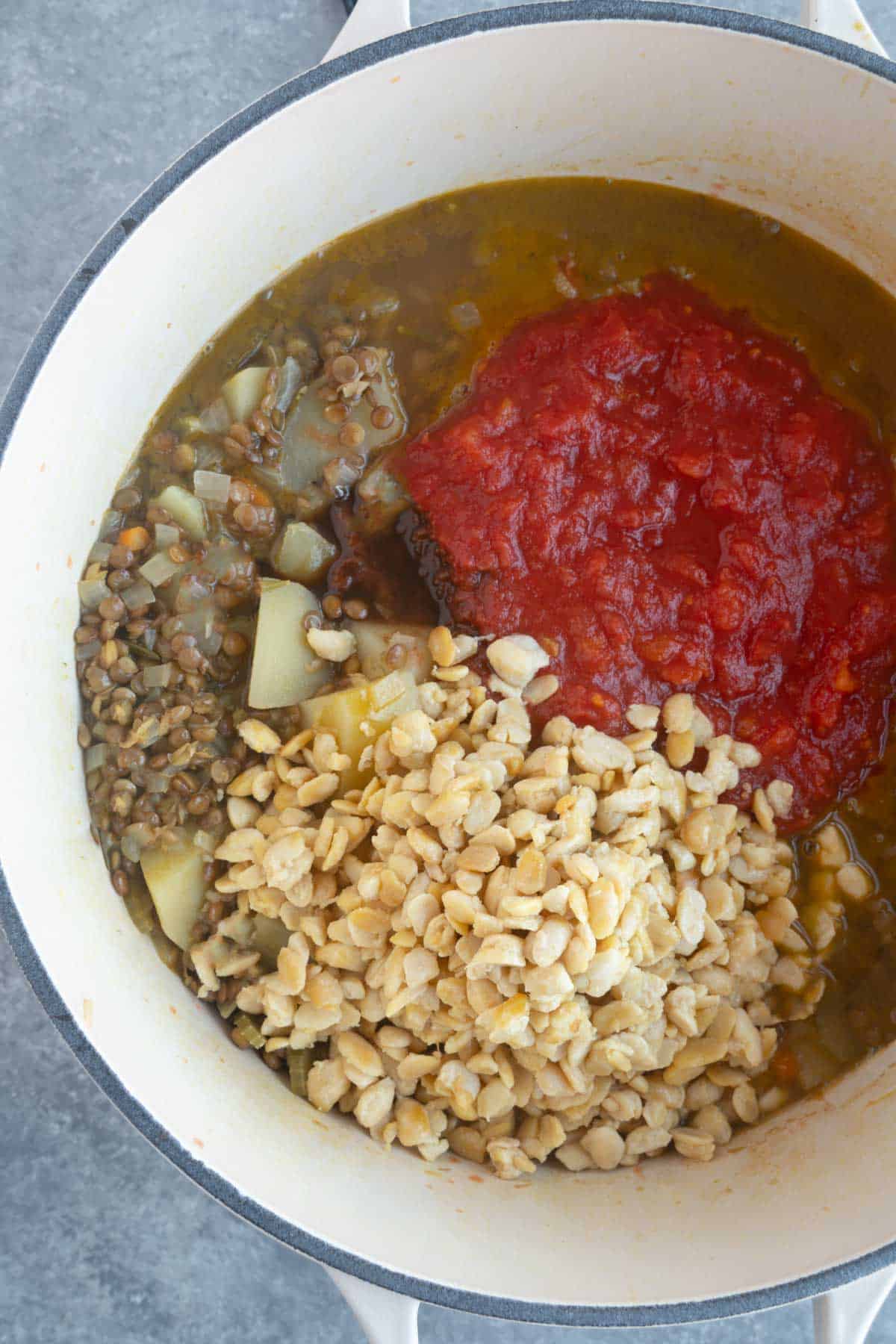 Crumbled tempeh, diced tomatoes, broth, lentils, and potatoes cooking in a white saucepan.