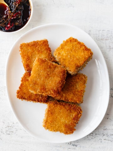 Crispy panko tofu squares on a white plate with a brown sauce and gold spoon.
