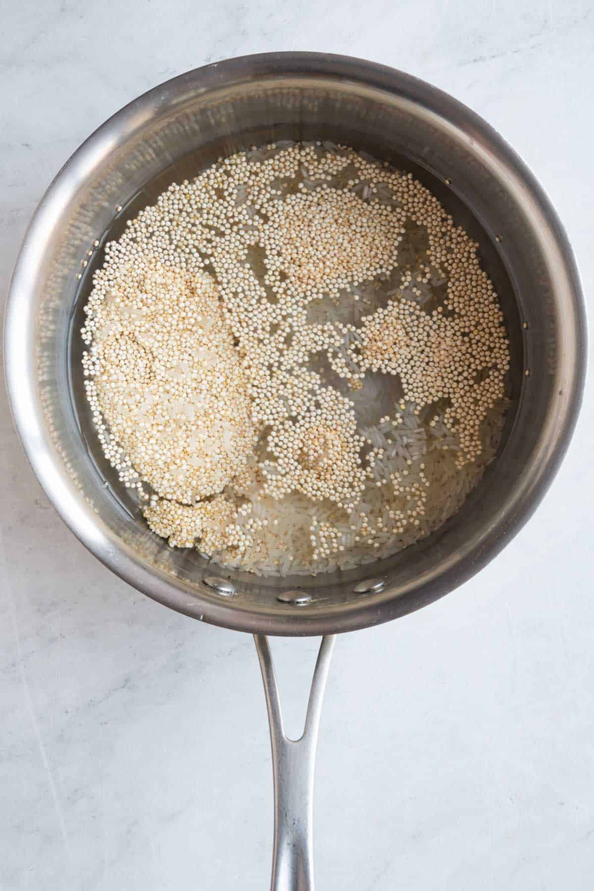 A stainless steel saucepan with water and dry quinoa floating on top with uncooked rice at the bottom.