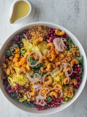 Roasted vegetable salad with quinoa, kale, squash, cashews, pickled onion, pomegranate, and cabbage in a white bowl with turmeric dressing.