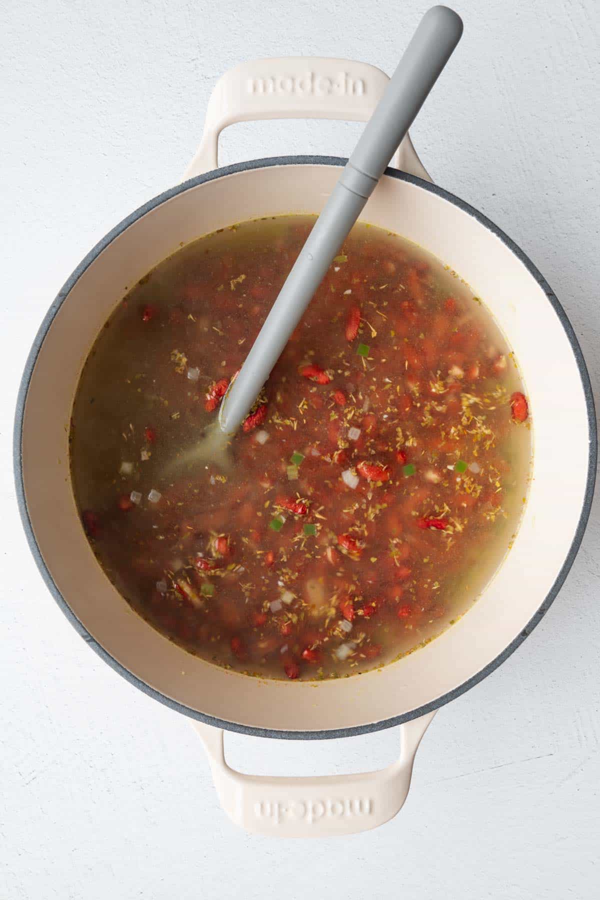 Red beans, herbs, and chopped veggies cooking in water in a white saucepan.