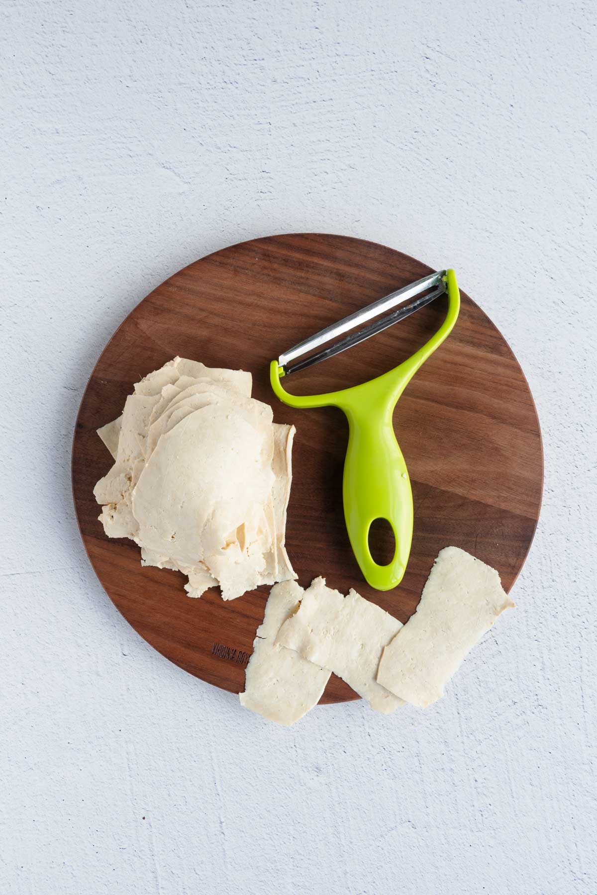 Grated tofu and a wide vegetable peeler on a round wooden cutting board.