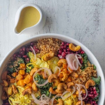 Yellow turmeric glow dressing in a white pouring bowl next to a roasted vegetable salad with cabbage, cashews, pomegranate, kale, and pickled shallots.