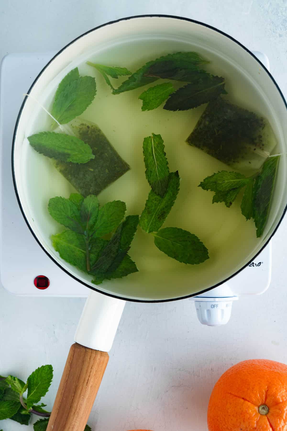 Green tea bags and mint leaves simmering in a white saucepan next to an orange, and fresh mint.