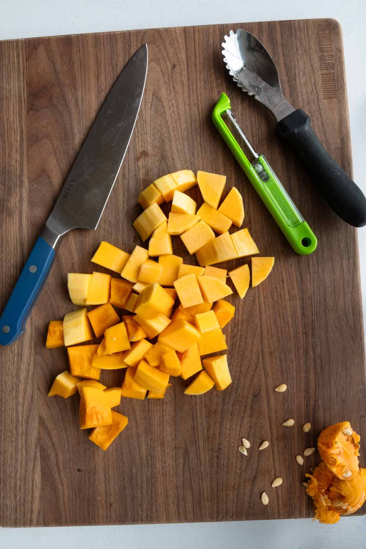 Cubed butternut squash on a wooden cutting board with a chef's knife, peeler, and scooping spoon.