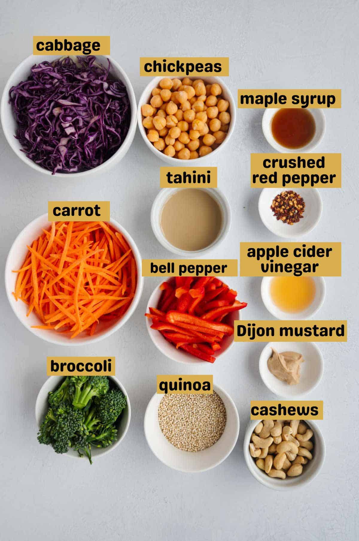 Shredded red cabbage, chickpeas, maple syrup, tahini, sliced carrots, sliced red bell pepper, apple cider vinegar, Dijon, cashews, quinoa, and broccoli in white bowls.