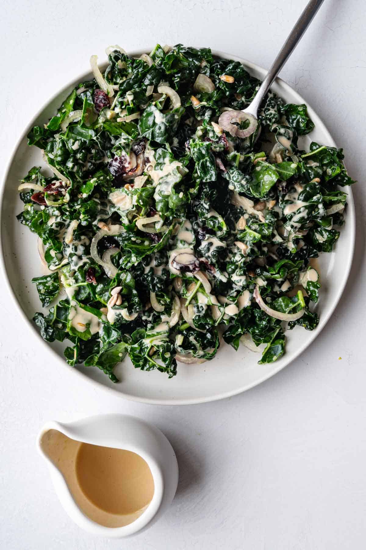 Kale salad with tahini dressing, sunflower seeds, shallots, and cranberries on a white plate with a silver fork.