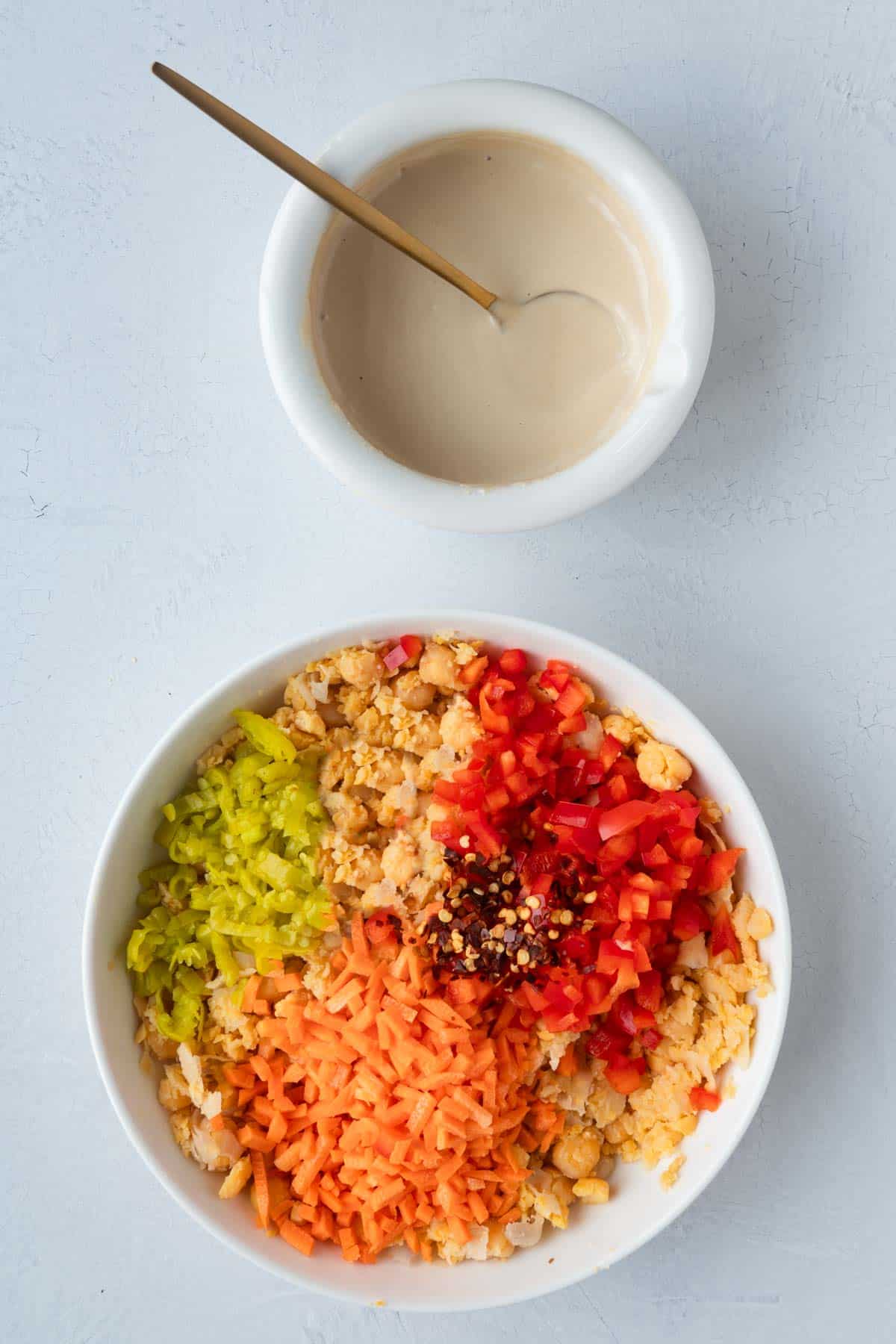 Chopped carrots, peppers, and pepperoncini, and red pepper flakes in a white bowl next to a smaller bowl with creamy tahini dressing.