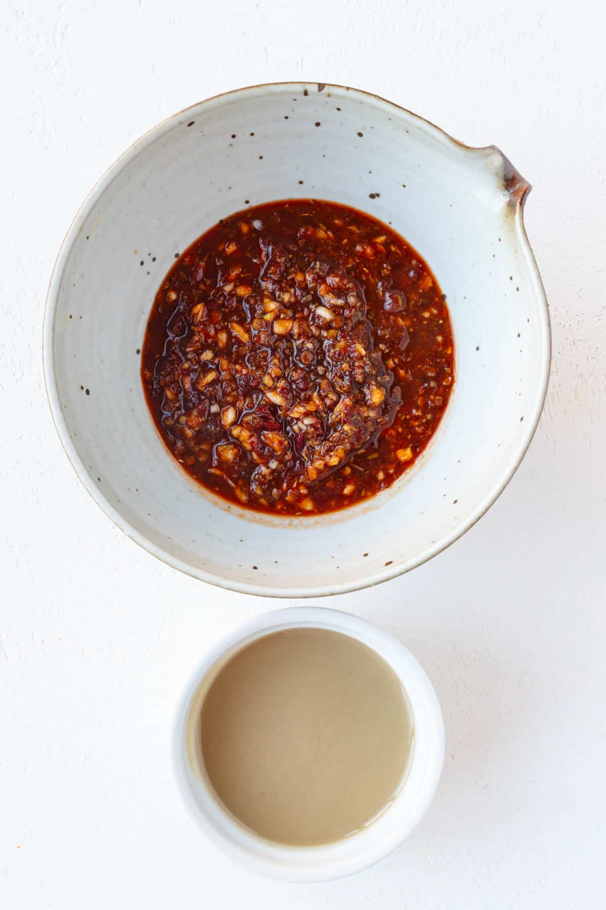 Red chipotle sauce with minced garlic next to a white bowl of tahini.