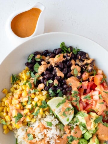 Baja sauce in a white bowl next to a Baja bowl with rice, black beans, corn, tomatoes, avocado, and lime.