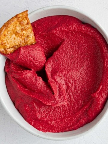 Pink beetroot hummus in a white bowl with one toasted pita wedge.