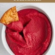 Pink beetroot hummus in a white bowl with one toasted pita wedge.