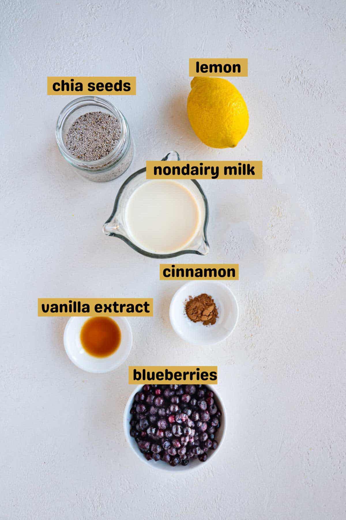 Chia seeds, lemon, soy milk, cinnamon, blueberries, and vanilla extract on a white board.