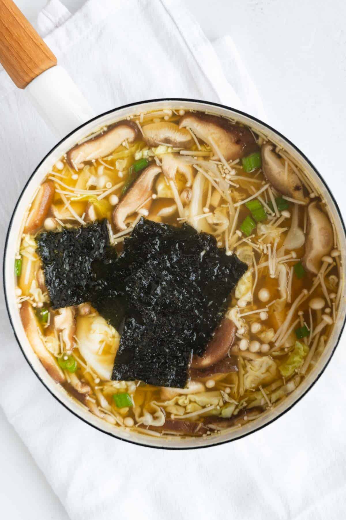 Miso soup with noodles, mushrooms, tofu, nori seaweed, and green onion in a white saucepan.