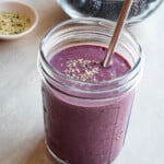 Mango blueberry chia smoothie (no bananas) in a glass with a metal straw with a bowl of berries and hemp hearts.