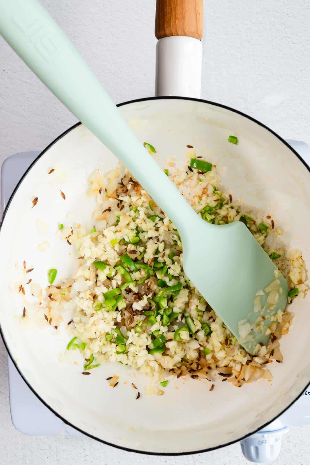 Cooked onion, garlic, green chili peppers, and cumin seeds in a white saucepan.