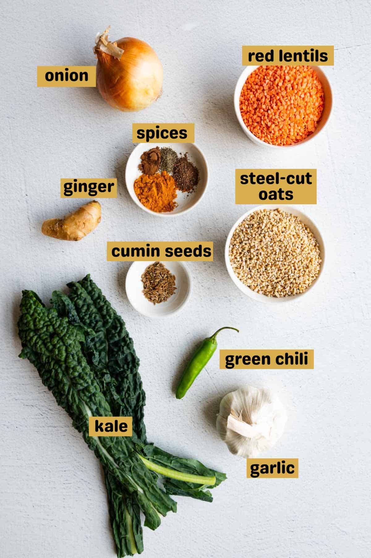Onion, red lentils, garlic, kale, green chili, cumin seeds, steel-cut oats, spices, and ginger on a white backdrop.